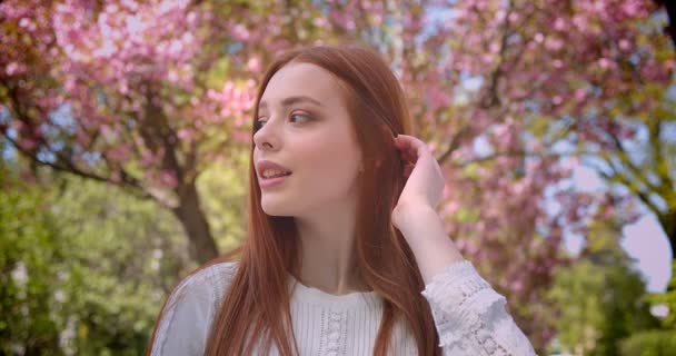 Charming ginger female student in white blouse watching dreamily into camera in pink floral garden. — Stock Video