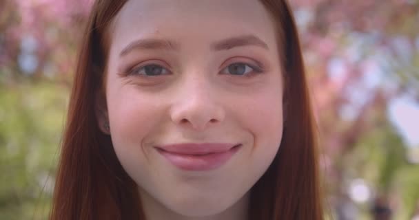 Close-up portrait of charming ginger female model watching dreamily and prettily into camera in pink floral garden. — Stock Video