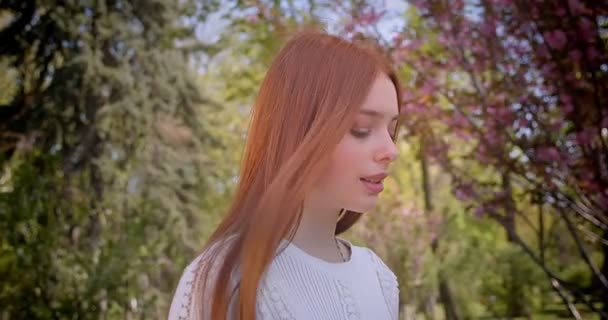 Ginger girl in white blouse walking along at pink floral garden and smiling into camera prettily. — Stock Video