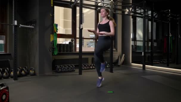 Молодая сконцентрированная спортсменка бегает с поднятыми коленями, а ее мобильный телефон лежит рядом в спортзале. . — стоковое видео
