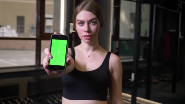 Sportsgirl mostrando smartphone com tela verde chromakey na câmera sendo sério e concentrado no ginásio . — Vídeo de Stock