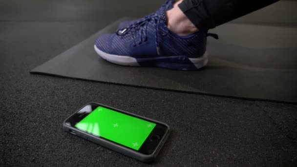 El teléfono móvil con pantalla verde cromakey que se encuentra cerca de las zapatillas de deporte en el karrimat en el gimnasio . — Vídeo de stock