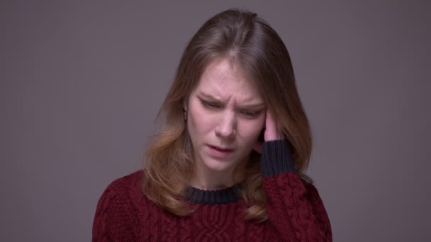 Portrait of depressed young female student having a migraine and scratching her head on gray background. — Stock Video