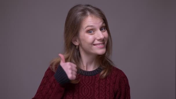 漂亮的年轻女学生把大拇指微笑着对着相机，以示喜欢和尊重灰色背景. — 图库视频影像