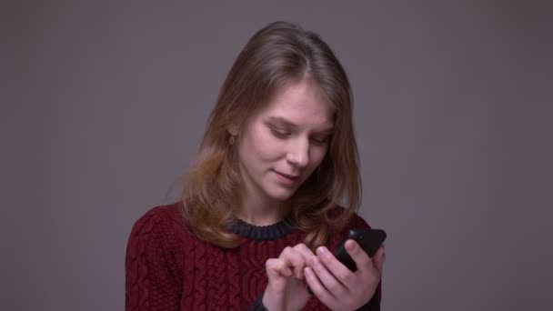 Jeune étudiante travaillant avec smartphone se tourne vers la caméra et sourit sur fond gris . — Video