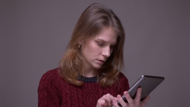 Joven estudiante femenina trabajando con la tableta siendo atento y concentrado se convierte en cámara sobre fondo gris . — Vídeo de stock