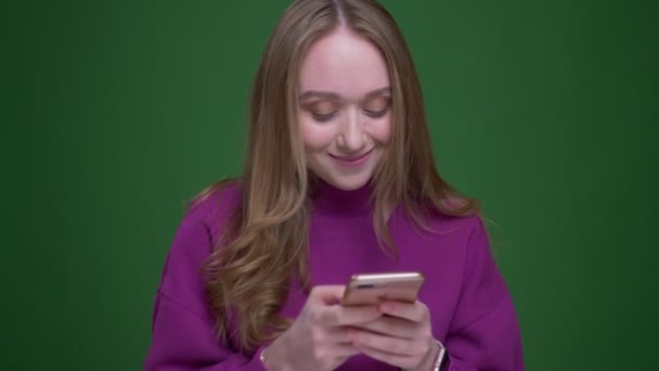 Güzel kadın zencefil öğrenci yeşil renk arka plan üzerinde kameraya dikkatli gülümsüyor telefon ile çalışan. — Stok video