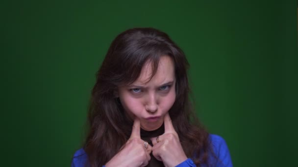 Young brunette female student make funny faces being joyful watching into camera on green background. — 비디오