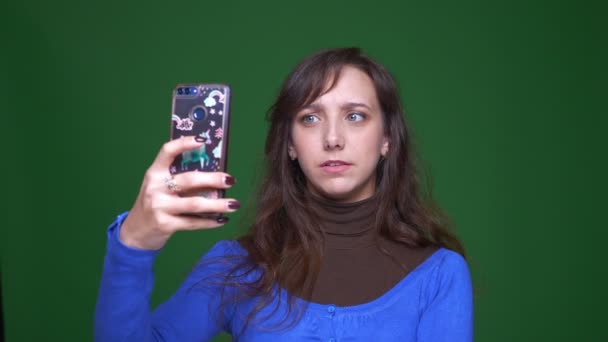 Ung brunette, kvinnelig student som tar selfiebilder smilende på smarttelefon på grønn bakgrunn . – stockvideo