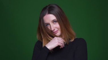Kestane rengi saçlı genç iş kadını saçlarıyla oynuyor ve yeşil arka planda kamerayla utangaç bir şekilde flört ediyor..