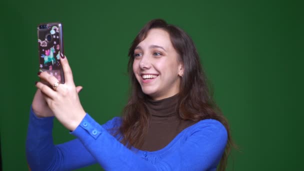 junge brünette Studentin spricht freudig im Videochat auf Smartphone auf grünem Hintergrund.