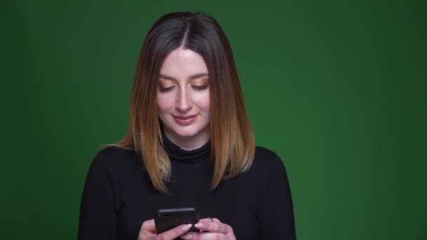 Joven empresaria con cabello castaño trabaja con el teléfono celular y reacciona emocionalmente sobre fondo verde . — Vídeo de stock