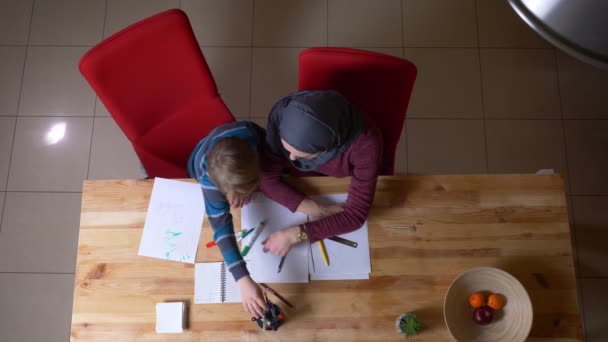 Kopfbild einer muslimischen Mutter im Hijab hilft ihrem Sohn, das Bild mit Filzstiften am Tisch zu zeichnen. — Stockvideo