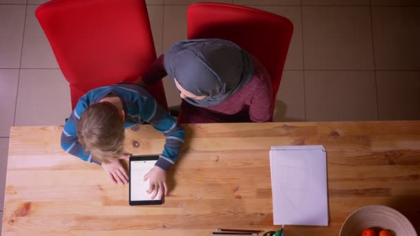 Top shot małego chłopca bawiącego się z tabletem i jego muzułmańską matką w hidżab obserwując swoją działalność siedzącą w pobliżu. — Wideo stockowe