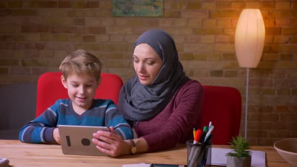 Retrato de menino pequeno e sua mãe muçulmana no hijab assistindo desenhos animados no tablet juntos e discutindo-os . — Vídeo de Stock