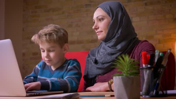 Kleine jongen spelen spelletjes op laptop en zijn moslim moeder in hijab observeren van zijn activiteit zitten in de buurt. — Stockvideo