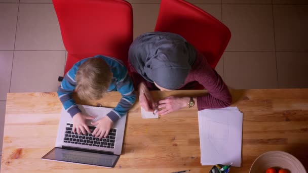 Top shot de niño pequeño escribiendo en el ordenador portátil y su madre musulmana en hijab escribir notas sentado cerca . — Vídeos de Stock