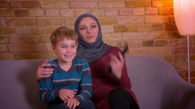 Küçük çocuk ve hijab onun Müslüman annesi portre sarılma ve evde kanepede otururken birlikte Tv izlerken.
