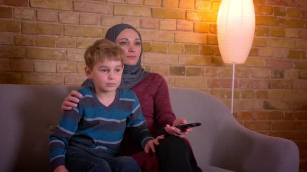 Portret van kleine jongen en zijn moslim moeder in hijab TV kijken samen zittend op de Bank thuis. — Stockvideo