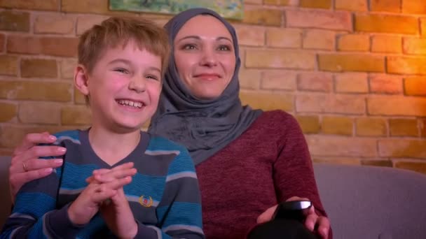 Портрет маленького мальчика и его мусульманской матери в хиджабе вспыхнул смехом смотря комедию по телевизору дома . — стоковое видео