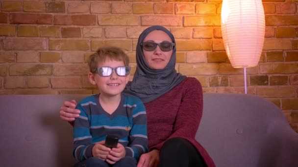 Retrato de niño pequeño y su madre musulmana en hijab en gafas 3D riendo mientras ve la comedia en la televisión en casa . — Vídeos de Stock