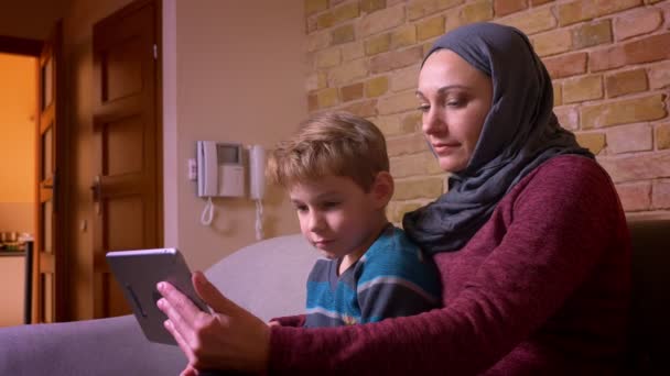 Retrato de un niño pequeño y su madre musulmana en hijab viendo películas en tabletas emocionados y divertidos en casa . — Vídeos de Stock