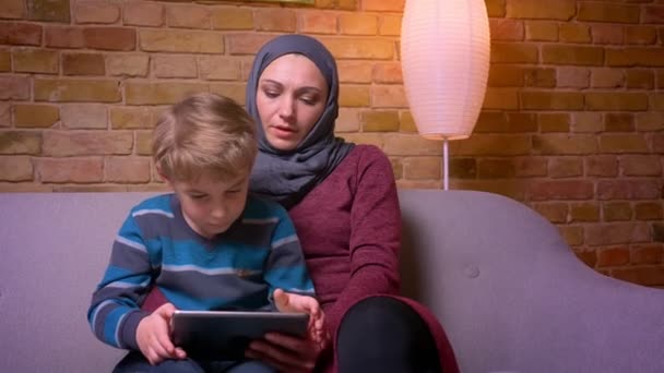 集中小男孩坐在他的穆斯林母亲在hijab玩平板电脑上游戏在家里的肖像. — 图库视频影像
