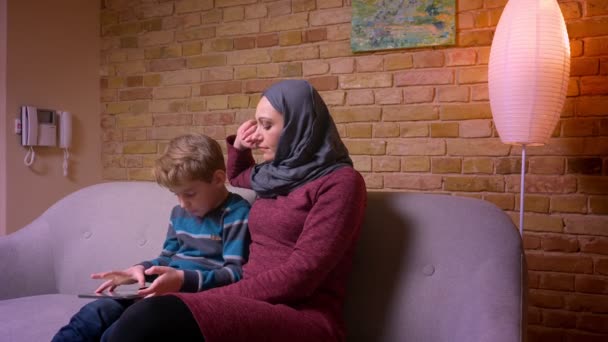 Konzentrierter kleiner Junge beim Spielen auf dem Tablet und seine muslimische Mutter im Hijab beobachten sein Treiben zu Hause. — Stockvideo