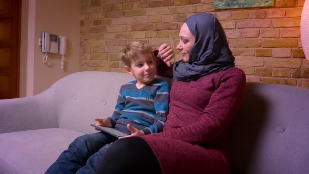 Συμπυκνωμένο μικρό αγόρι παίζοντας παιχνίδι στο tablet και τη μουσουλμανική μητέρα του σε χιτζάμπ τον χαϊδεύουν τρυφερά στο σπίτι. — Αρχείο Βίντεο