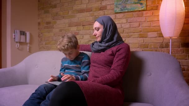 Menino divertido jogando jogo no tablet e sua mãe muçulmana no hijab observa sua atividade em casa . — Vídeo de Stock