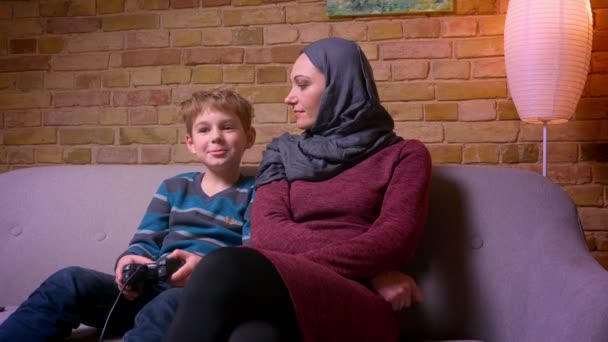 Концентрированный маленький мальчик играет в видеоигру с джойстиком, а его мусульманская мать в хиджабе помогает ему выиграть дома . — стоковое видео