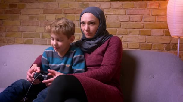 Geconcentreerde kleine jongen en zijn moslim moeder in hijab spelen video game met joystick samen thuis. — Stockvideo