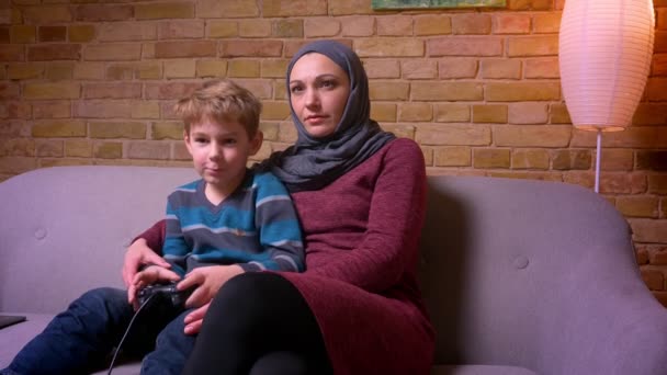 집중된 작은 소년 재생 비디오 게임 과 그의 무슬림 어머니 에 히잡 시도 에 선택 용 조이스틱 에 시도 재생. — 비디오