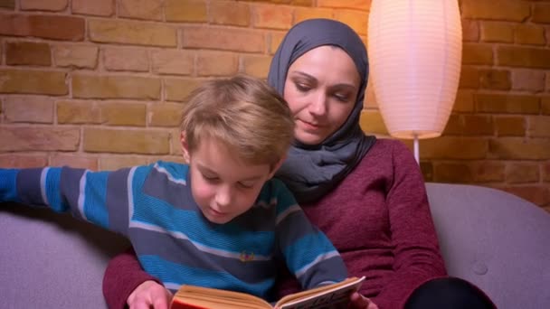 Nahaufnahme eines konzentrierten kleinen Jungen und seiner muslimischen Mutter im Hijab, die zu Hause aufmerksam Buch lesen. — Stockvideo