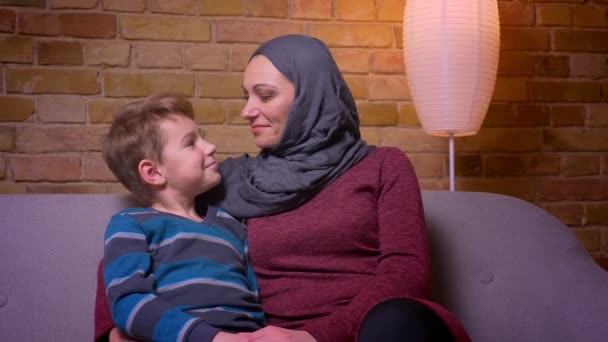Close-up tiro de menino pequeno e sua mãe muçulmana no hijab sorrindo na câmera inclinar a cabeça um para o outro em casa . — Vídeo de Stock