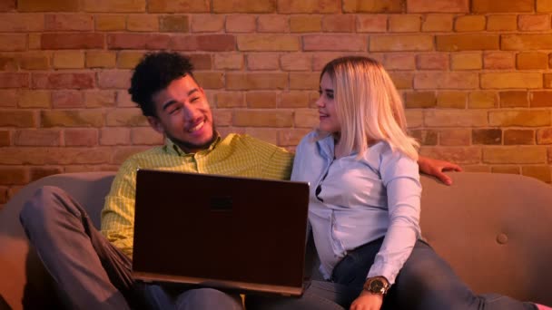 Young african guy and caucasian girl sitting on sofa and watching into laptop being joyful and relaxed at home. — ストック動画