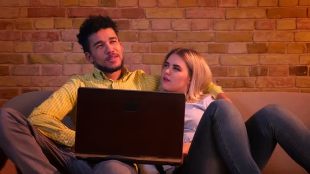 Young african guy and caucasian girl sitting on sofa with laptop and talking with each other joyfully at home. — ストック動画