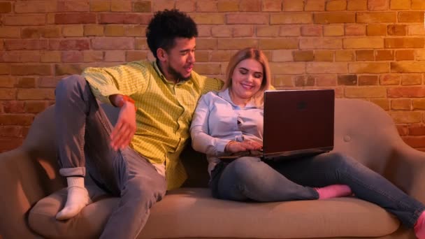 African guy and caucasian girl sitting on sofa with laptop and flirting with each other joyfully at home. — Stock Video