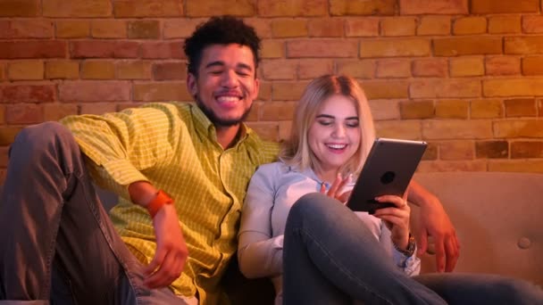 African guy and caucasian girl sitting on sofa with tablet and laughing joyfully at home. — Stock Video