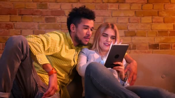 Africano cara e caucasiano menina sentado no sofá assistindo juntos em tablet e discutir em casa . — Vídeo de Stock