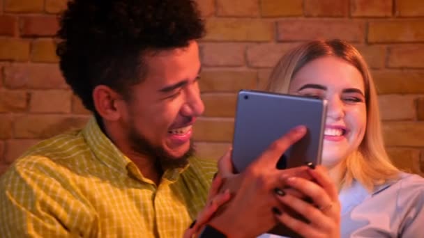 African guy and caucasian girl sitting on sofa talking funny selfie-photos on tablet being joyful and happy at home. — Stock Video