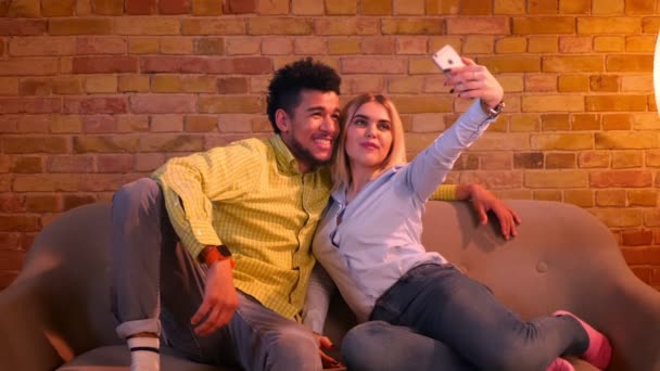 African guy and caucasian girl sitting on sofa making selfie-photos on phone being joyful and pretty together at home. — Stock Video