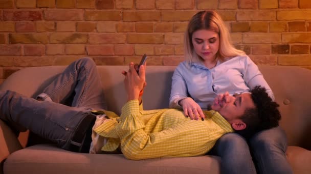 スマートフォンを持つアフリカの男は、彼の白人のガールフレンドの足に横たわり、自宅の雰囲気の中で自分撮り写真を作ります. — ストック動画
