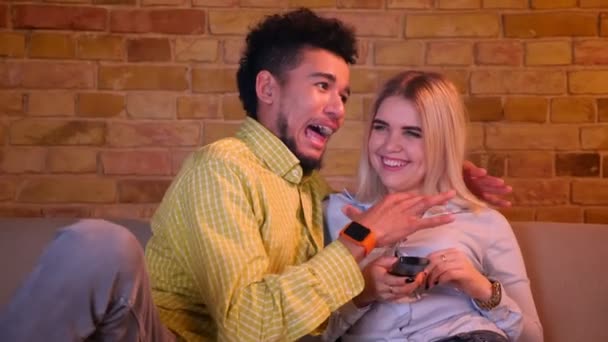 Young african guy strangles his blonde caucasian girlfriend watching TV in cozy home atmosphere. — ストック動画