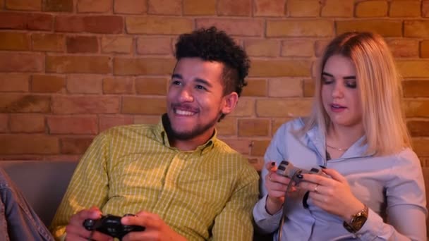 Jonge Afrikaanse jongen met zijn blonde blanke vriendin spelen videogame met joysticks in gezellige huiselijke sfeer. — Stockvideo