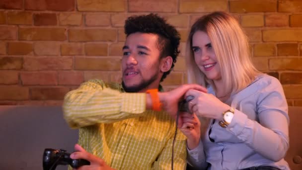 Afrikaner mit seiner blonden kaukasischen Freundin spielt Videospiel mit Steuerknüppel aufmerksam in gemütlichem Heim. — Stockvideo