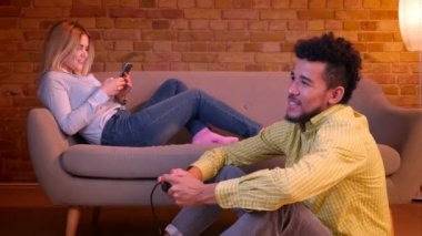 Afrikalı adam joystick ve kafkas kız ile video oyunu oynarken katta oturur rahat evde kanepeüzerinde akıllı telefon ile çalışır.