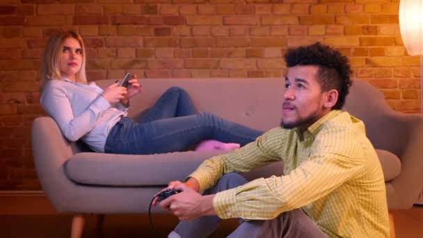 Africano cara se senta no chão jogando videogame com joystick e caucasiano menina trabalha com smartphone no sofá em casa aconchegante . — Vídeo de Stock