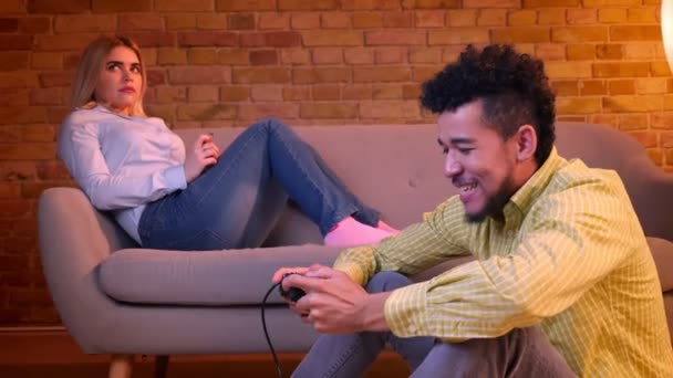Africano chico se sienta en piso jugando videojuego alegremente y caucásico chica con smartphone en acogedor hogar . — Vídeo de stock