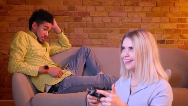 Blonde girl plays videogame on floor attentively and african boyfriend feeling depressed and lonely on background. — Stock Video